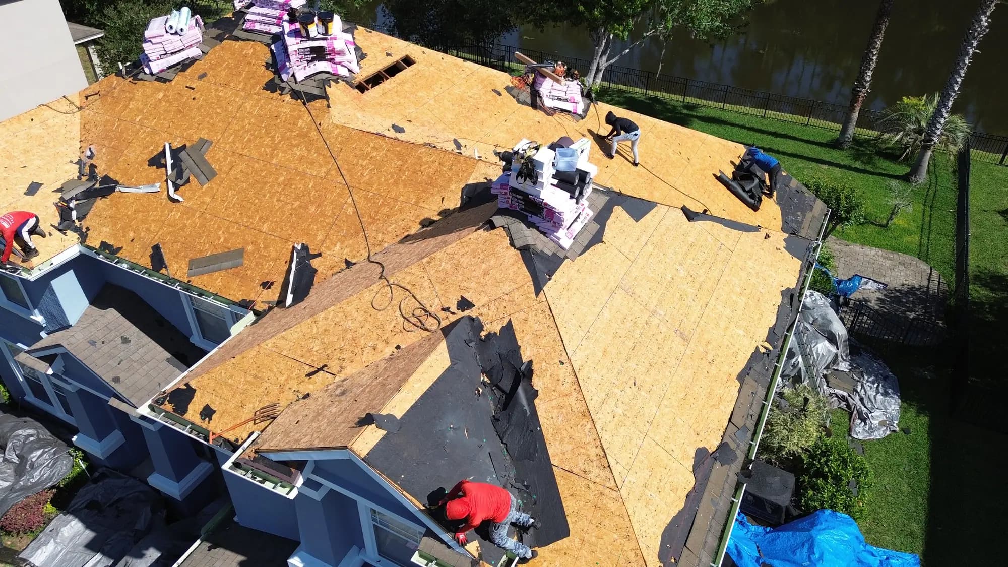 Aerial view of roofing materials and crew distributed across a stripped roof deck beside a pond