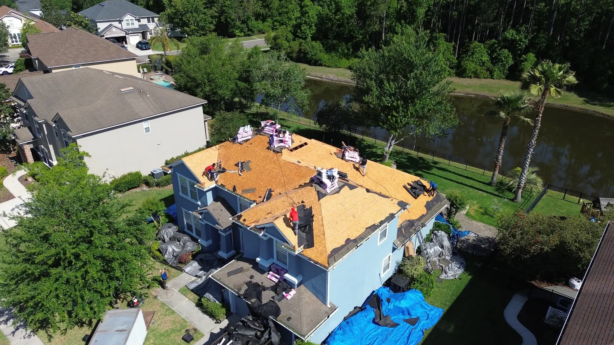 Roofing crew working across exposed roof decking on a blue two-storey home beside a pond