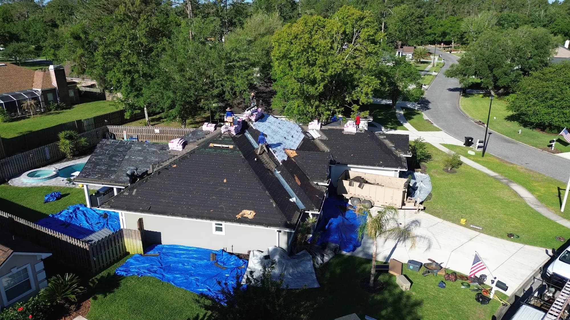Single-storey Florida home with synthetic underlayment being installed over the roof deck, dump trailer in the driveway and tarps protecting the pool and landscaping