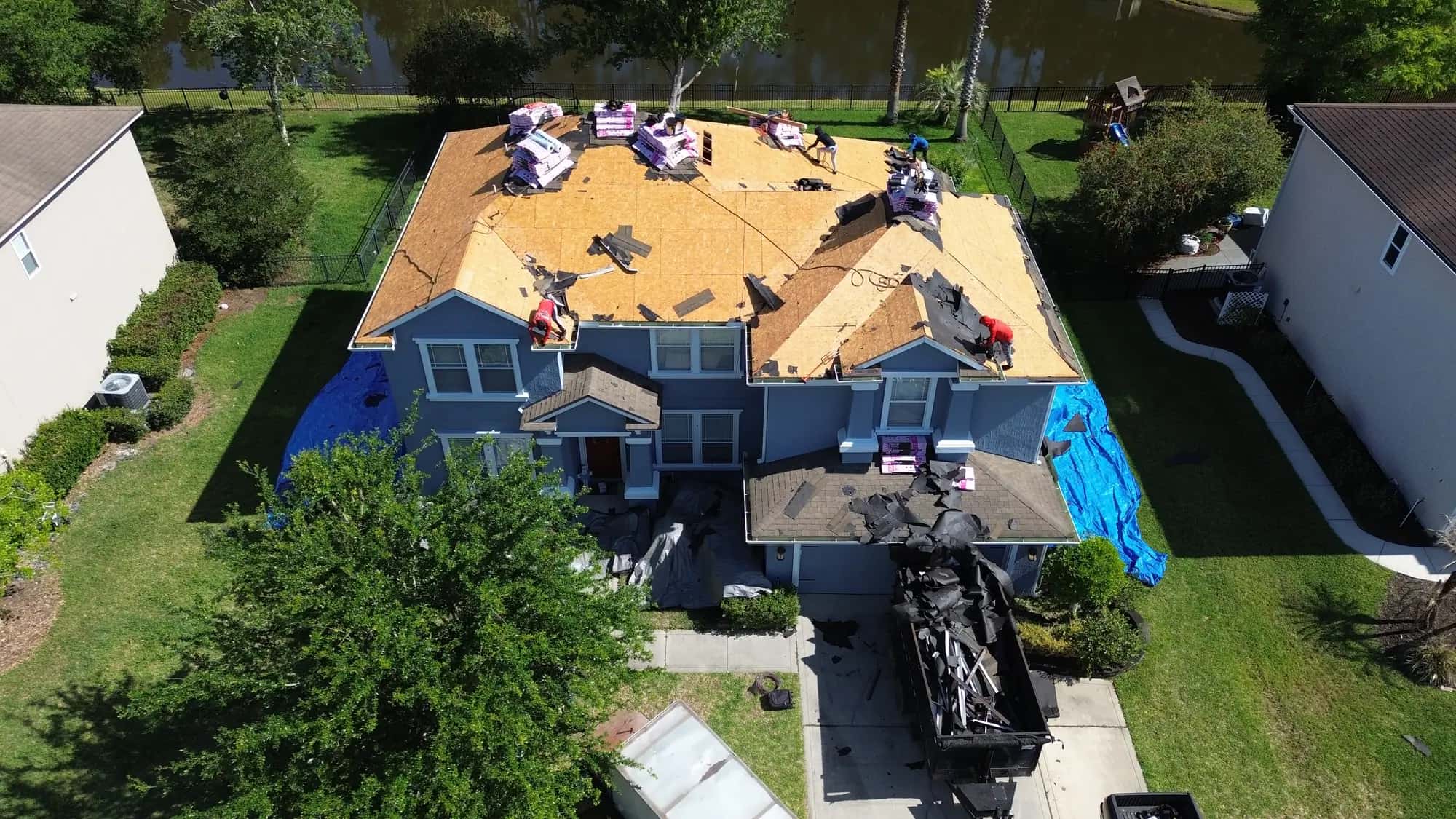 Aerial view of a two-storey Jacksonville home stripped to bare decking, with a Diversity Contracting crew working and a dump trailer of old shingles in the driveway
