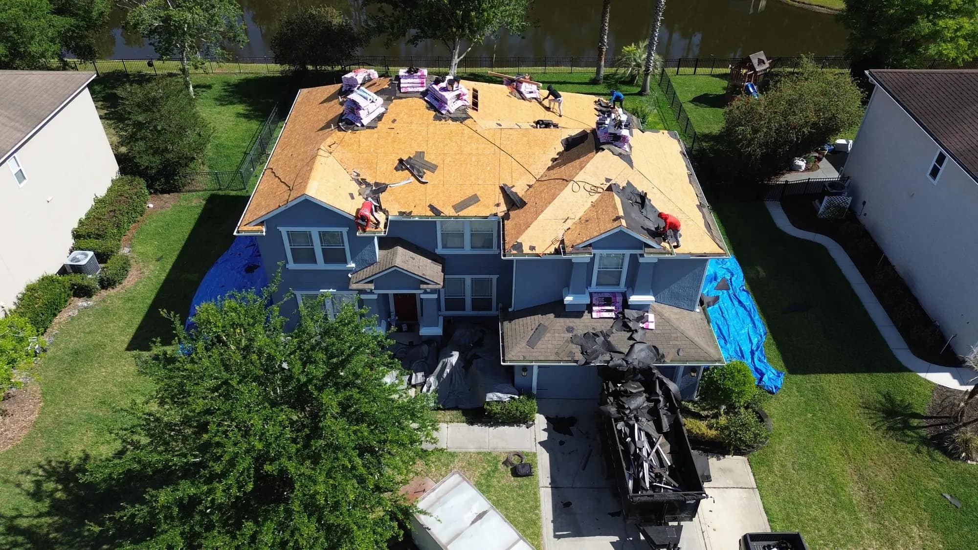 Aerial view of a two-storey Jacksonville home stripped to bare decking, with a Diversity Contracting crew working and a dump trailer of old shingles in the driveway